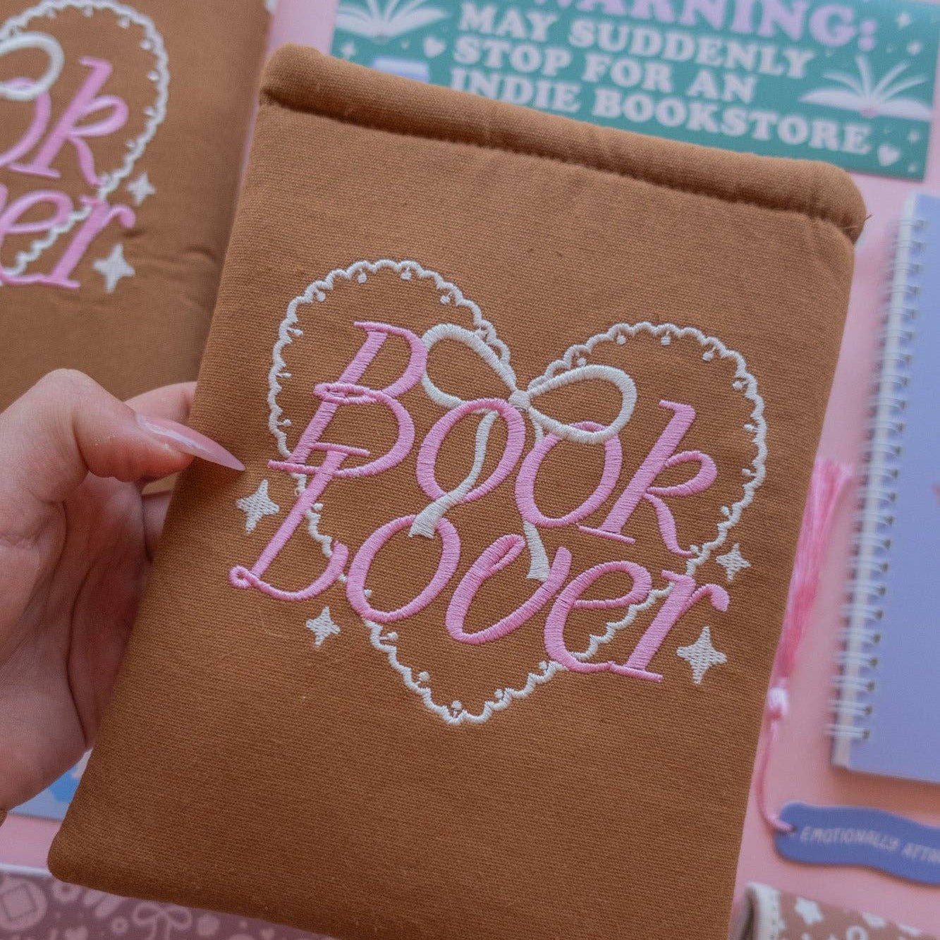 Brown notebook with 'Book Lover' design held by a hand, with other notebooks and stationery items in the background.