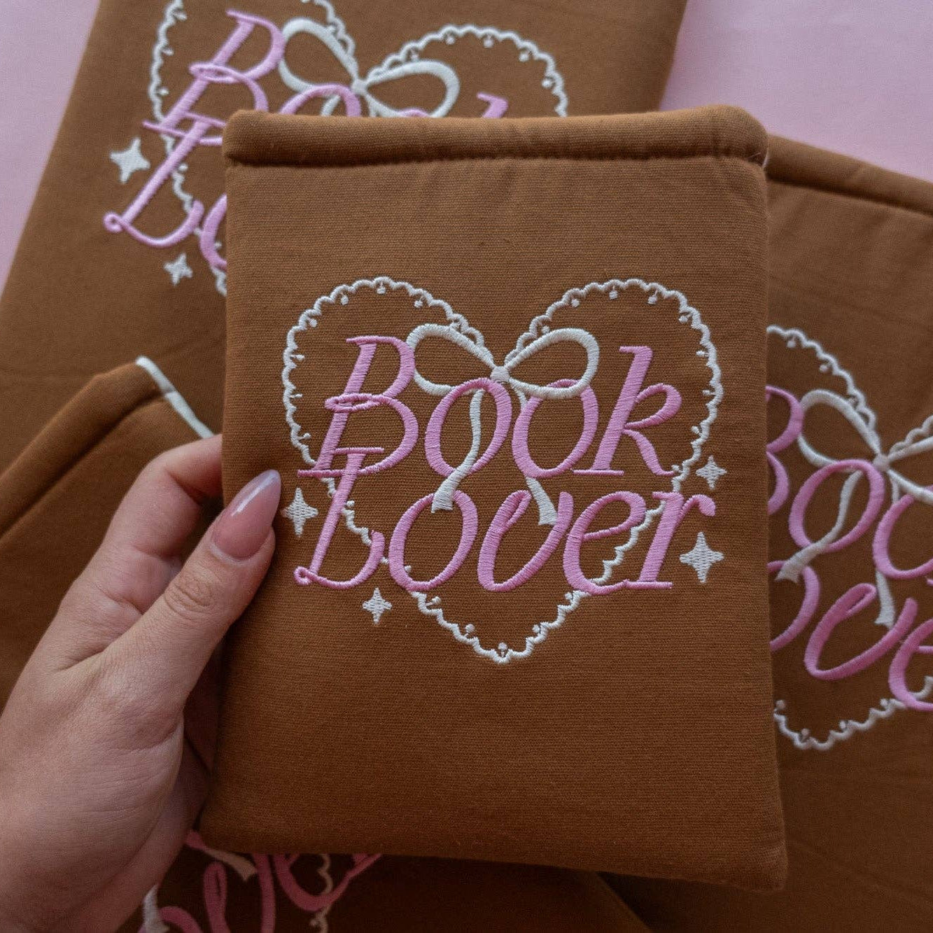 Brown pillow with 'Book Lover' embroidery held by a hand against a pink background