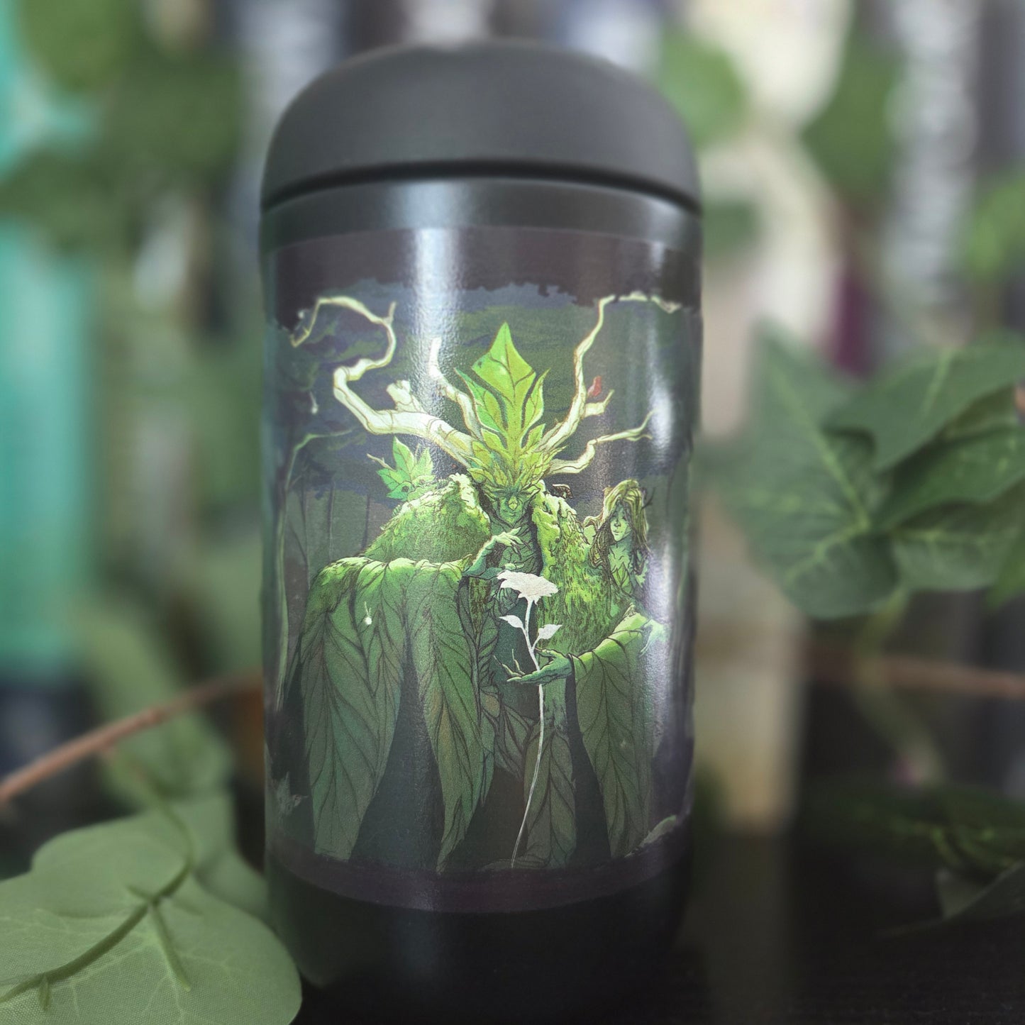 A black insulated tumbler with a matching lid, featuring a detailed graphic of a green mossy plant with glowing roots and a small white flower. The tumbler is photographed outdoors with blurred green leaves in the background.